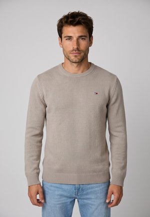 Mann mit kurzen braunen Haaren, der einen beigen Strickpullover und hellblaue Jeans trägt, steht vor einem einfarbigen grauen Hintergrund.