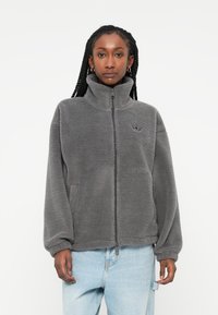 Giacca in pile grigia con colletto alto, zip completa, spalle scese e un piccolo logo sul petto. Tessuto morbido con una vestibilità rilassata.
