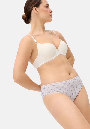 Mujer con sujetador acolchado blanco y ropa interior gris claro con pequeños estampados florales azules, posando con las manos detrás de la espalda.