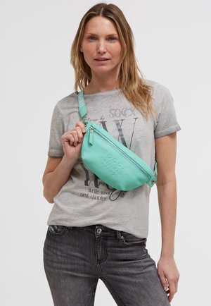 Woman wearing a gray graphic t-shirt and dark jeans holding a mint green leather crossbody bag with zipper and embossed text.