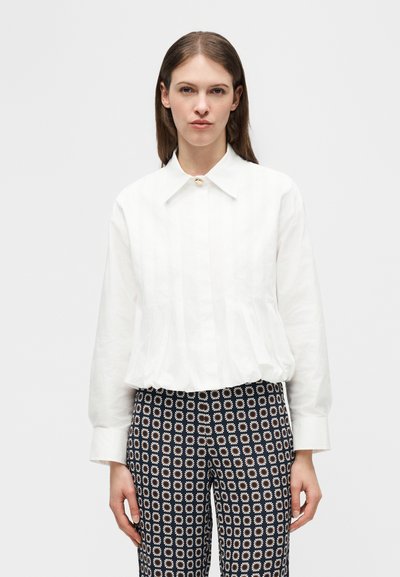 Femme aux cheveux bruns lisses portant un chemisier blanc à manches longues et un pantalon noir à motifs de marguerites blanches, debout devant un fond uni.