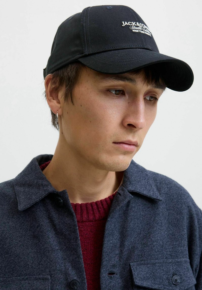 Jaunas vyras vilki juodą Jack & Jones kepurę, tamsiai pilką švarką su knopkėmis ir bordo spalvos megztinį, žiūri žemyn su neutralia išraiška.
