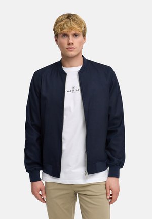 Junger Mann mit blondem Haar, der eine marineblaue Zip-Jacke über einem weißen T-Shirt mit Grafik und beigen Hosen trägt, steht vor einem einfarbigen Hintergrund.