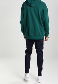 Grön huvtröja med avslappnad passform, i kombination med marinblå joggers med vita sidoränder, och vita träningsskor.