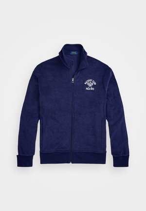 Chaqueta de forro polar azul marino con cremallera y cuello alzado, que presenta el texto blanco "Polo NYC" y un gráfico de raqueta de tenis en el lado izquierdo del pecho. Textura suave.