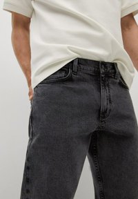 Jeans in denim grigio scuro con taglio dritto, caratterizzati da una sottile trama. Abbinati a una polo bianca con texture, mani nelle tasche.