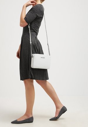 Vrouw in een donkergrijze knielange jurk met glinsterende grijze ballerina's en een witte schoudertas met een kettingriem.