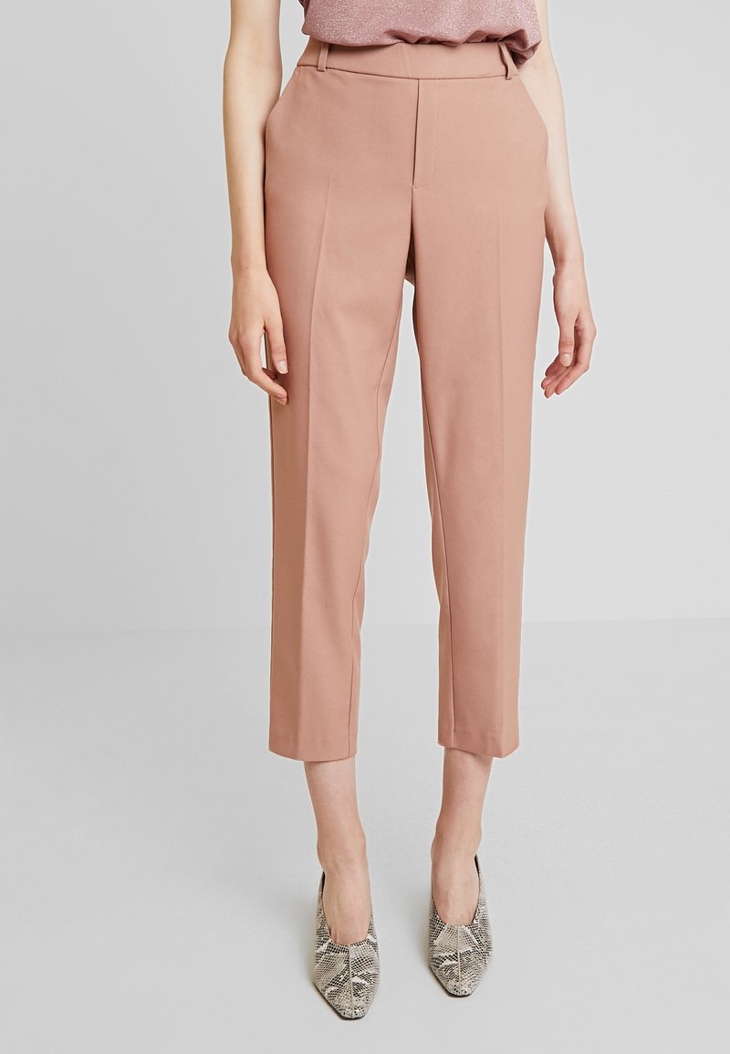 Femme portant un pantalon court taille haute rose poudré associé à des escarpins pointus à imprimé peau de serpent sur un fond uni.