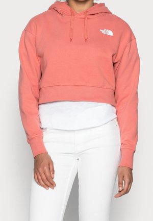 Korte roze capuchontrui gemaakt van zacht materiaal. Heeft geribde manchetten en zoom, met een wit logo op de linkerborst. Witte gelaagde blouse eronder.