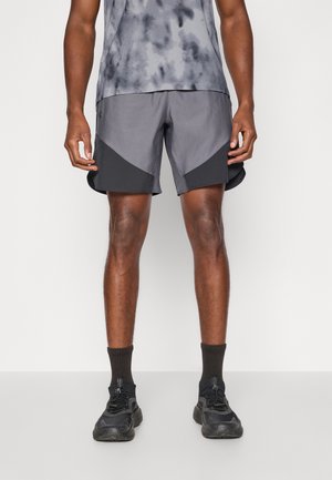 Homme portant un short de sport gris et noir, des baskets noires, des chaussettes noires et un débardeur gris à motifs, debout devant un fond uni.