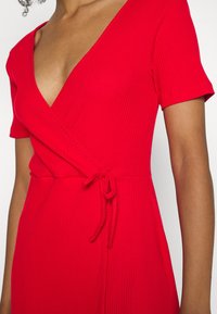 Czerwona, żeberkowana sukienka kopertowa z krótkimi rękawami, dekoltem w kształcie litery V i wiązaniem w talii. Gładka tekstura i dopasowany krój podkreślają design.