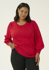 Donna con capelli castani di media lunghezza che indossa un maglione rosso con maniche a sbuffo e pantaloni neri, in piedi davanti a uno sfondo chiaro e semplice.