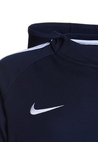 Felpa blu navy con collo alto e dettagli bianchi, caratterizzata da un piccolo logo Nike argentato sulla parte anteriore. Realizzata in tessuto morbido e elasticizzato.