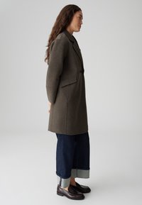 Vrouw in profiel, gekleed in een donkerbruine wollen jas, spijkerbroek met brede manchet en donkerbruine leren loafers, tegen een grijze achtergrond.