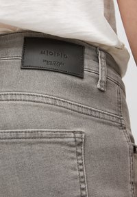 Jeans de denim gris con una superficie texturizada, que presentan un parche de cuero negro etiquetado "Marc O'Polo Denim" en la cinturilla.