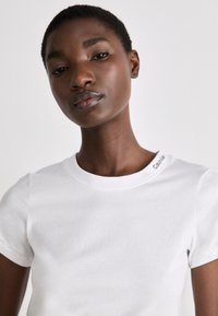 T-shirt bianco in cotone con collo rotondo e maniche corte. Presenta un piccolo logo sulla spalla in un colore a contrasto. Texture liscia.