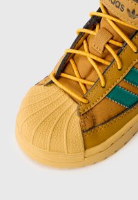 Sárga sportcipő texturált gumitalppal, szövet felsőrésszel és zöld bőrcsíkokkal. Sárga fűzőkkel és texturált talppal rendelkezik.