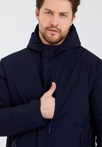 Veste bleu marine avec capuche, fermeture éclair à l'avant et deux poches latérales zippées. Présente une texture lisse et mate ainsi qu'un design minimaliste.