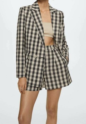 Femme portant un blazer à carreaux noir et blanc et un short taille haute assorti avec un crop top beige, une main dans la poche du short.