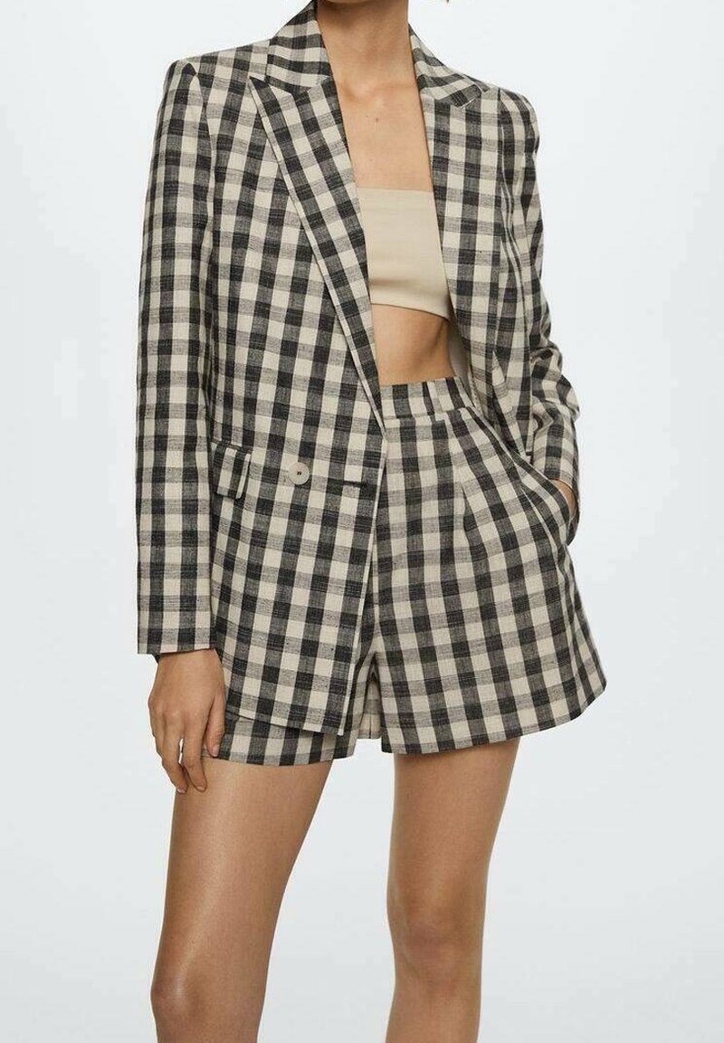 Femme portant un blazer à carreaux noir et blanc et un short taille haute assorti avec un crop top beige, une main dans la poche du short.
