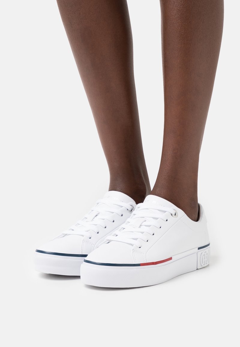 Personne portant des baskets basses blanches avec un détail de bandes bleues et rouges sur des semelles blanches, debout sur un fond clair uni.