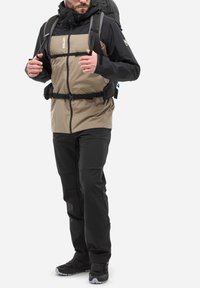 Veste d'extérieur beige et noire avec fermeture éclair à l'avant, taille ajustable et poches latérales, associée à un pantalon noir et des chaussures de randonnée.