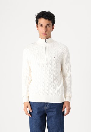 Cremfarbener Strickpullover mit hohem Kragen und Reißverschluss. Weiche Textur, gerippte Bündchen und Saum. Kombiniert mit Jeans.