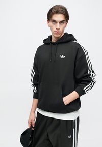 Čierna mikina s kapucňou a predným vreckom, biele logo Adidas na hrudi a tri biele pruhy po stranách rukávov. Materiál vyzerá mäkko a textúrovo.