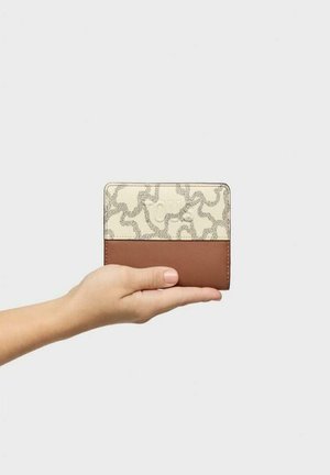 Billetera bicolor con una parte superior crema con un patrón texturizado y una sección inferior de cuero marrón, sostenida en una mano contra un fondo neutro.