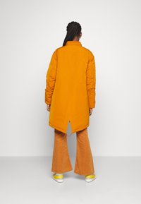 Personne avec des cheveux tressés portant une longue veste matelassée orange, un pantalon flare en velours côtelé orange et des baskets jaunes, debout devant un fond uni.