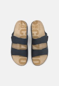 Mornarsko modre natikače z dvema prilagodljivima trakoma, mehka bež podloga z vzorcem blagovne znamke in gladka tekstura, podobna usnju.