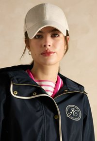 Jūras spēku ūdensnecaurlaidīga jaka ar zelta aparatūru, baltām malām un logo uzšuvumu. Zem tās, rozā un baltas svītru džemperis un bēšs cepure.