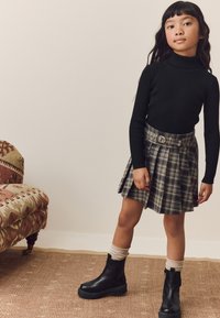 Black ribbed turtleneck, plaid pleated skirt with belt, black ankle boots, cream knitted socks. Geometric patterns on sofa, textured rug.