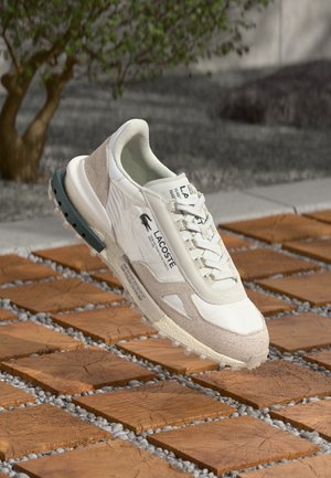 Sneaker Lacoste alb și bej deschis, cu un superior din textură combinată, călcâi întărit și o talpă din cauciuc cu crampoane pentru aderență.