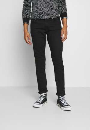 Personne portant un pantalon noir slim, des baskets montantes noires et blanches, et un haut noir à manches longues avec un texte imprimé blanc.