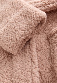 Tissu doux, brun clair, texturé bouclé, plié serré, ressemblant à du polaire ou du sherpa, montrant une surface détaillée et des coutures.