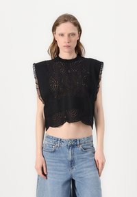Top corto nero con orlo scalloped, caratterizzato da motivi circolari ricamati e dettagli in pizzo sulle spalle. Indossato con jeans azzurri chiari.