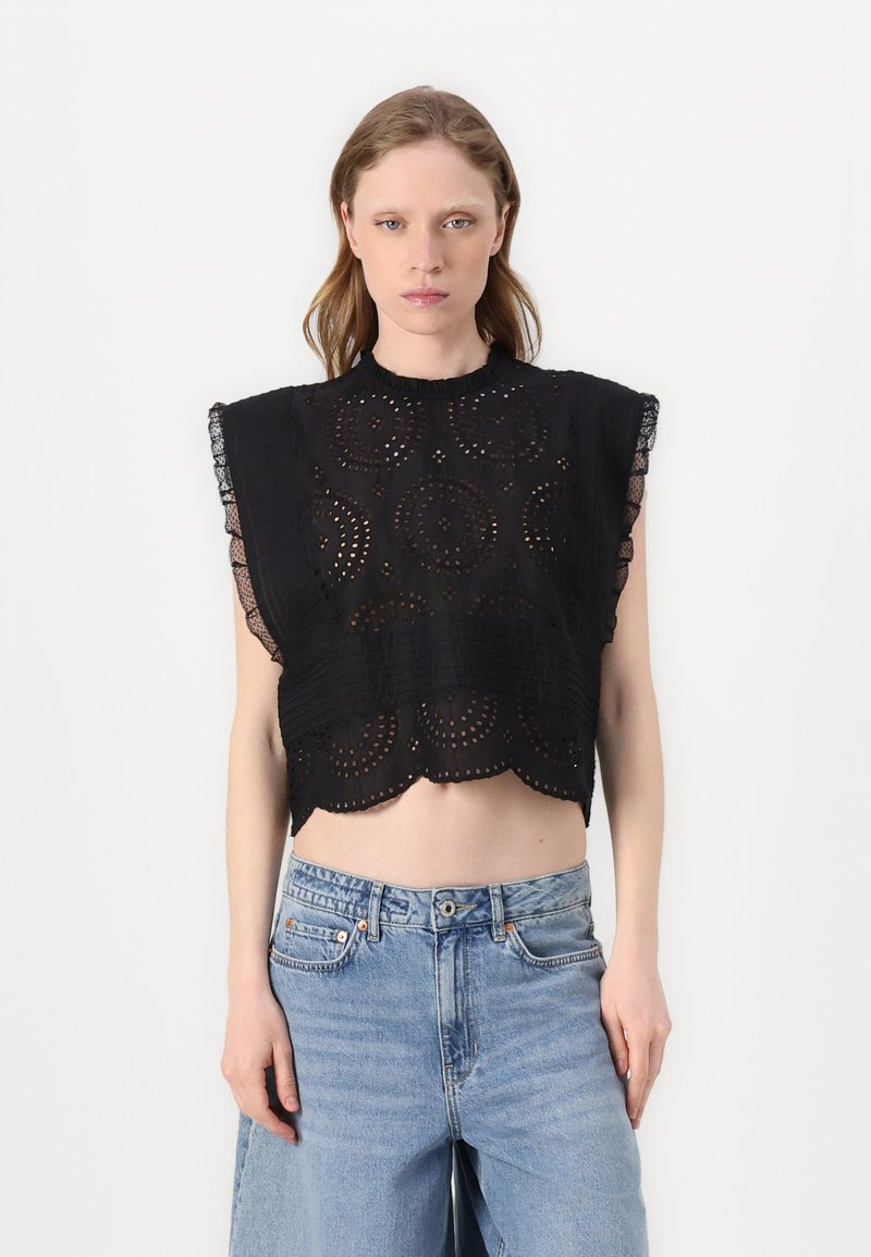 Top corto nero con orlo scalloped, caratterizzato da motivi circolari ricamati e dettagli in pizzo sulle spalle. Indossato con jeans azzurri chiari.