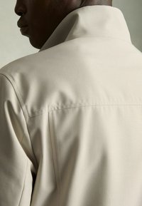 Cămașă bej deschis cu guler înalt, textură fină și detalii de cusătură vizibile. Prezintă un design curat, ajustat, fără embellishments vizibile.