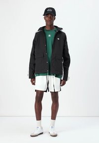 Μαύρο αδιάβροχο μπουφάν με λευκές ρίγες, πράσινη μπλούζα από κάτω, λευκά σορτς και αθλητικά παπούτσια. Τα αξεσουάρ περιλαμβάνουν ένα μαύρο καπέλο με κείμενο.