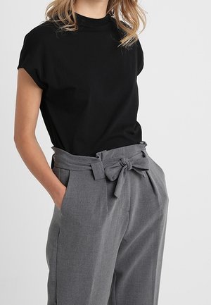 Vrouw die een zwart top met korte mouwen draagt, ingestopt in een grijze, hooggetailleerde broek met een stoffen riem die is vastgeknoopt, met haar handen in de zakken.