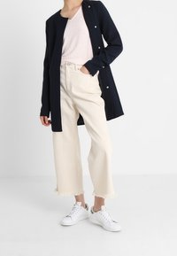 Mörkblå lång kofta över en ljusrosa topp, kombinerad med vita vida byxor med fransiga muddar och vita sneakers med gröna detaljer.