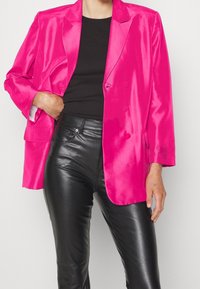 Różowy satynowy blazer w intensywnym odcieniu z podwójnym zapięciem, klapami, kieszenią na piersi, zestawiony z dopasowanymi czarnymi skórzanymi spodniami.