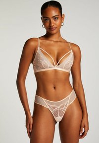 Nude bralette met kantafwerking en V-hals ontwerp. Bijpassende doorzichtige string met kantdetails en uitsnijdingen aan de zijkanten.