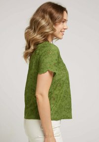 Blouse verte brodée avec des motifs floraux, des manches à volants et une coupe décontractée. Le tissu est léger avec des découpes.