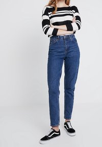 Džínové kalhoty s vysokým pasem v modré barvě, se zužujícími nohavicemi, nošené s černobílým pruhovaným topem s dlouhým rukávem a černými teniskami.