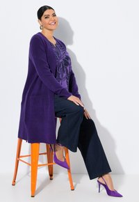 Lila stickad cardigan över en lila blommig topp, kombinerad med mörka jeans och lila stilettklackar, sittande på en orange pall.