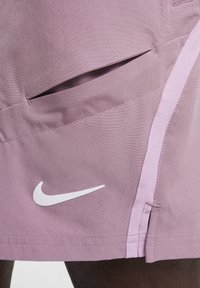 Nike-short in lichtpaars stof, met een zijzak en een witte logo-accent. Soepele textuur met contrasterende lichtroze naden.