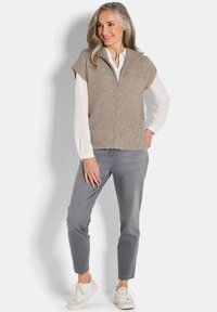 Gilet sans manches beige avec un col haut et une fermeture éclair sur le devant, porté sur une chemise blanche et un pantalon gris, assorti à des baskets de couleur claire.