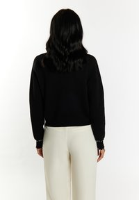 Schwarzer Pullover mit langen Ärmeln, der mit Perlenakzenten an den Bündchen versehen ist, kombiniert mit cremefarbenen, hoch taillierten Hosen.